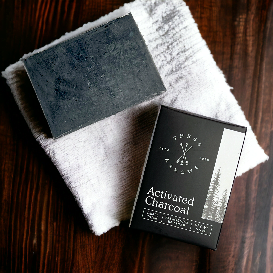 A box of charcoal bar soap next to a bar of charcoal bar soap on a dark wooden surface